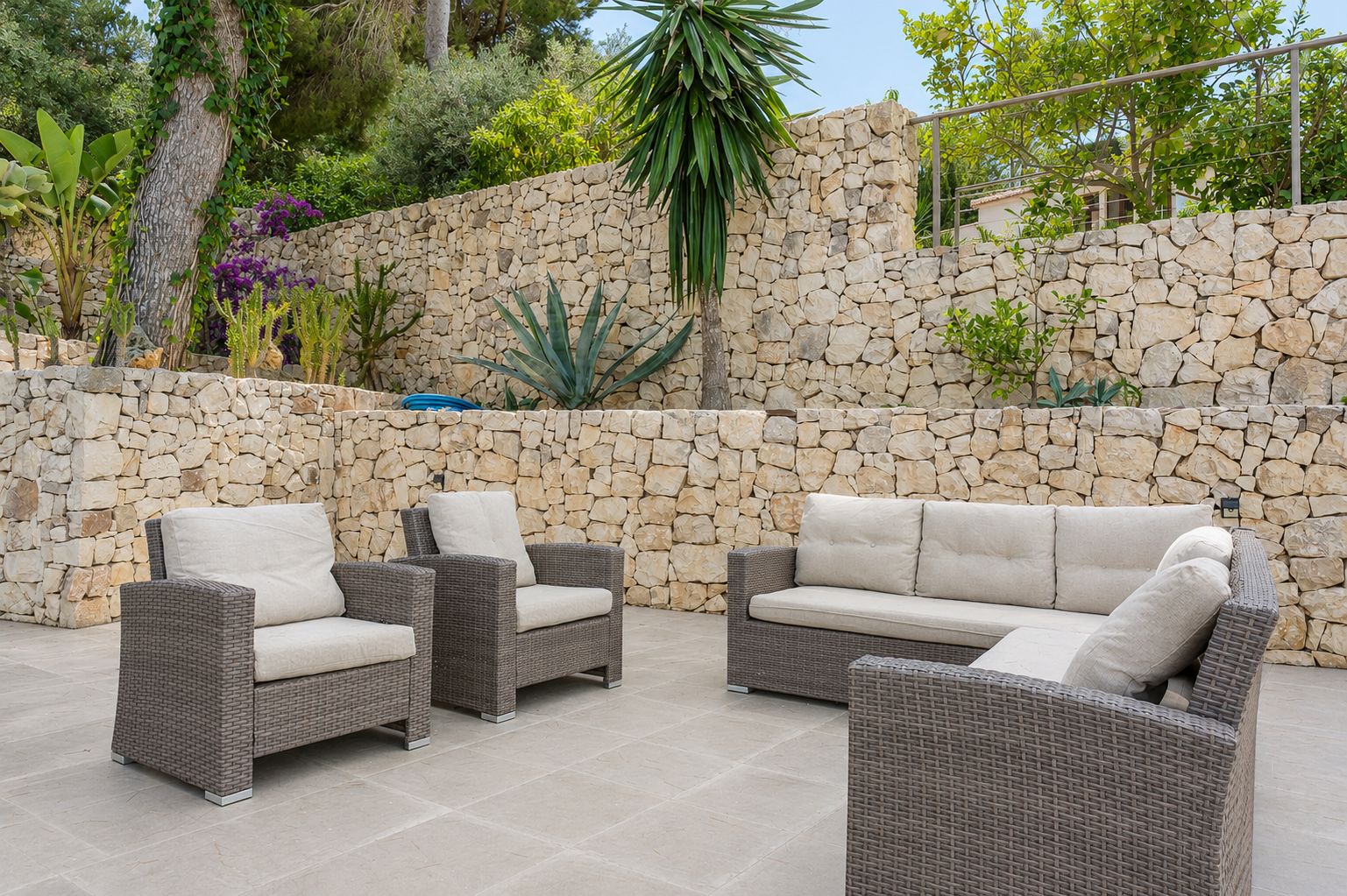 Grande terrasse extérieure avec mobilier en rotin et coussins clairs, encadrée par des murs en pierre sèche étagés et une végétation méditerranéenne luxuriante.