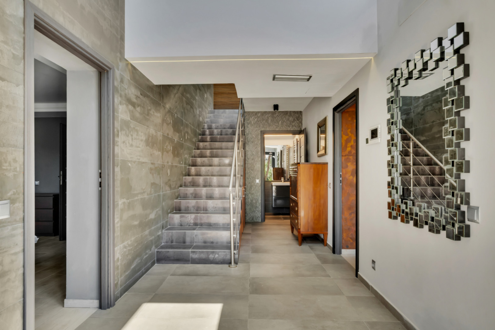 Hall d'entrée spacieux avec un escalier moderne revêtu de carreaux effet béton et une rampe en acier inoxydable. Le sol est carrelé de gris et un miroir modulaire décore le mur.