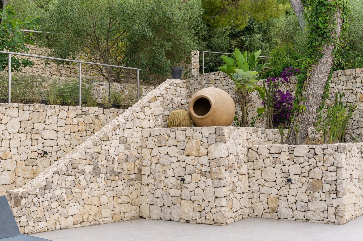 Jardin en terrasses avec murs en pierres sèches, escaliers intégrés et flore méditerranéenne variée. Une grande jarre décorative et des cactus soulignent l'esthétique naturelle et rustique.