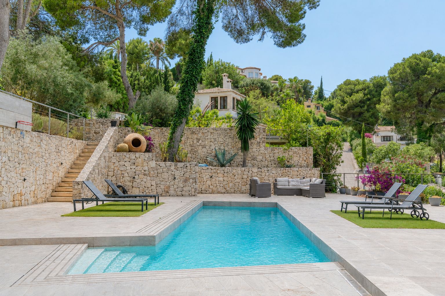 Piscine privée rectangulaire avec escaliers intégrés, bordée de dalles claires et de gazon synthétique avec transats. Murs en pierre naturelle et végétation méditerranéenne en terrasses.