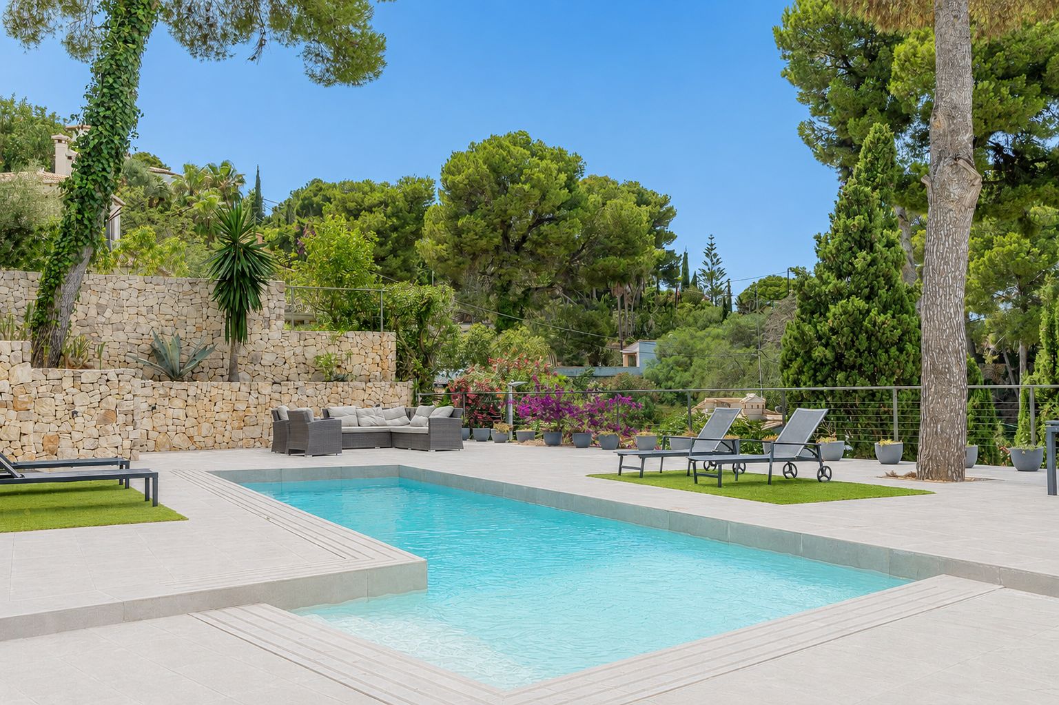 Piscine privée rectangulaire bordée d'une vaste terrasse carrelée claire, avec salon de jardin et mur en pierre naturelle au cœur d'une végétation luxuriante.
