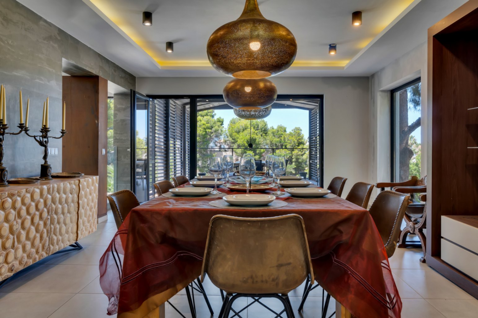 Salle à manger élégante avec des suspensions en bronze ouvragées, un buffet en bois texturé et de grandes fenêtres offrant une vue paisible sur la verdure luxuriante.