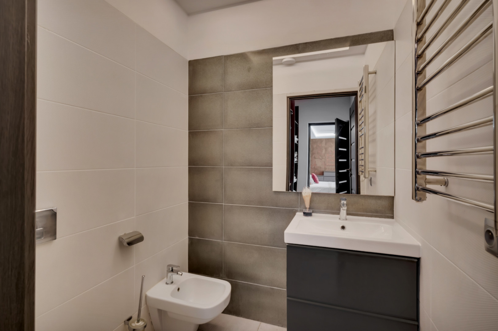 Salle de bain contemporaine avec meuble-lavabo gris foncé, vasque blanche, bidet et sèche-serviettes chromé. Les murs associent des carreaux clairs texturés et des carreaux foncés lisses.