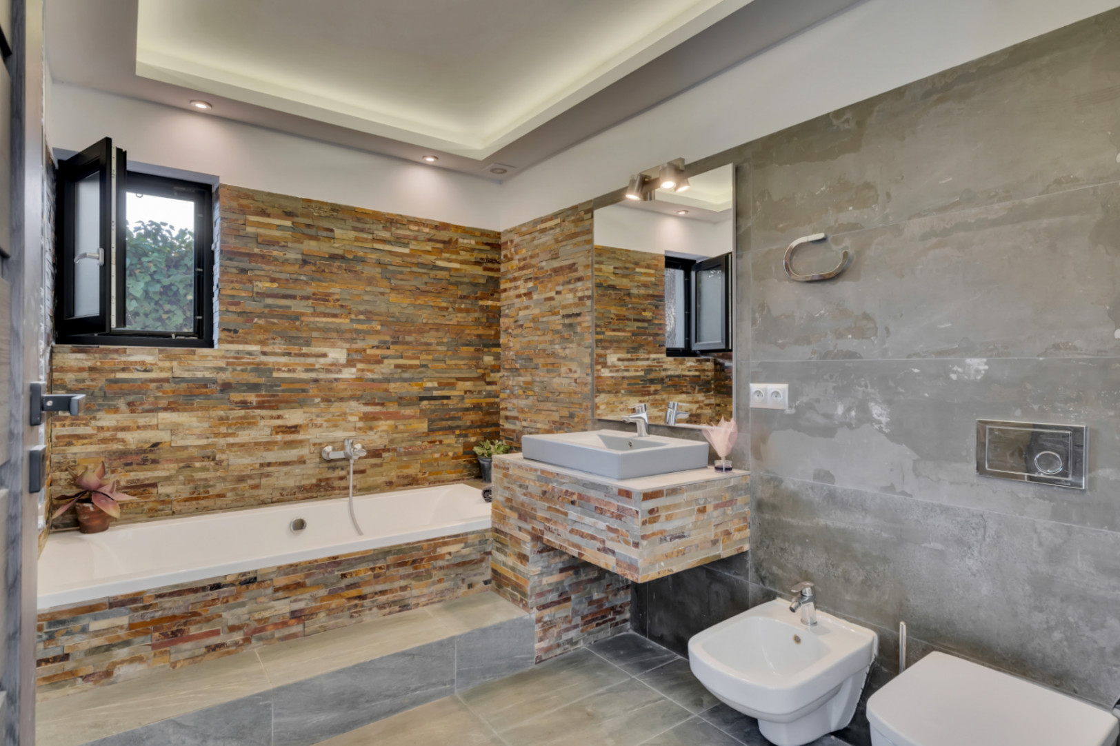 Salle de bain moderne avec baignoire et meuble-lavabo suspendu, tous deux revêtus de pierre empilée multicolore. De grands carreaux gris habillent murs et sol, éclairage encastré.