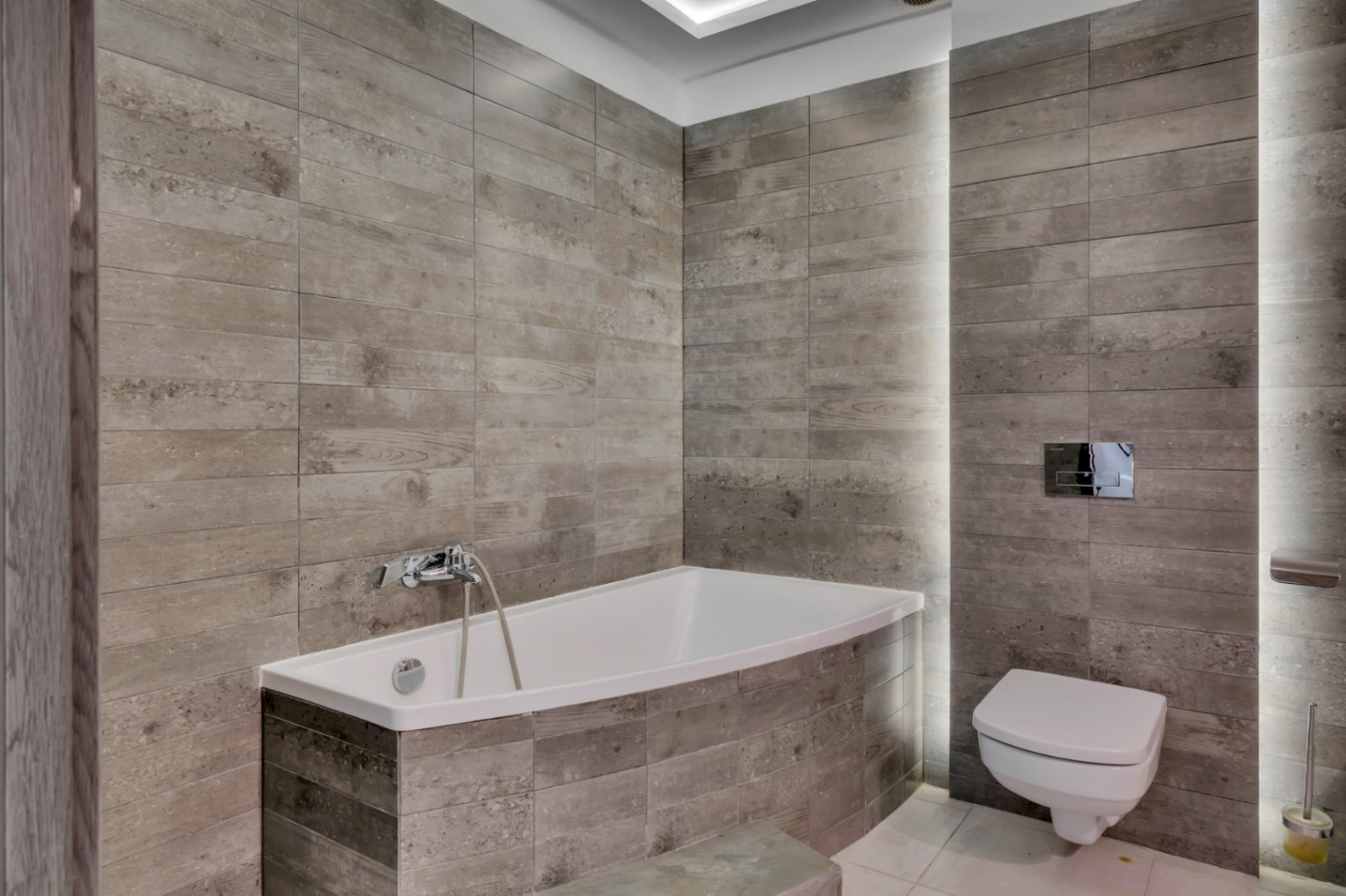 Salle de bain moderne avec baignoire intégrée et WC suspendu, revêtue de carreaux céramiques gris texturés et éclairage LED vertical intégré.