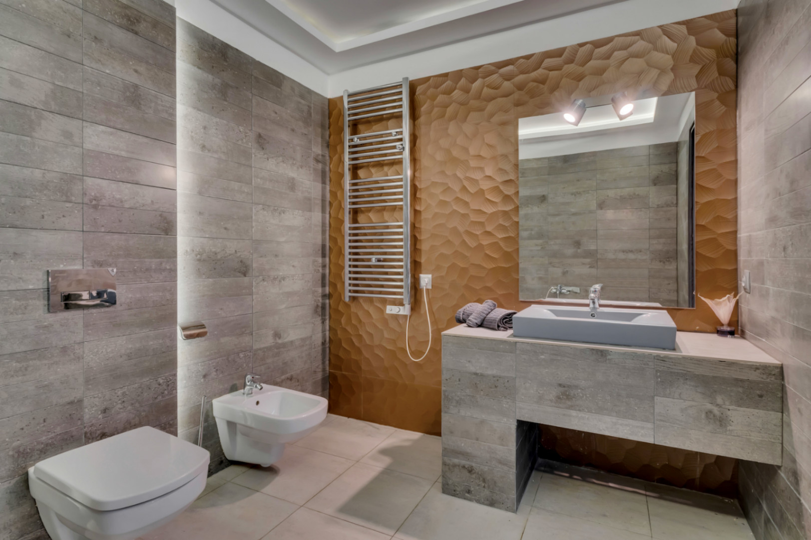 Salle de bain moderne avec carrelage mural gris texturé, un mur d'accent 3D géométrique bronze et un meuble-lavabo suspendu. Comprend toilettes et bidet, ainsi qu'un sèche-serviettes.