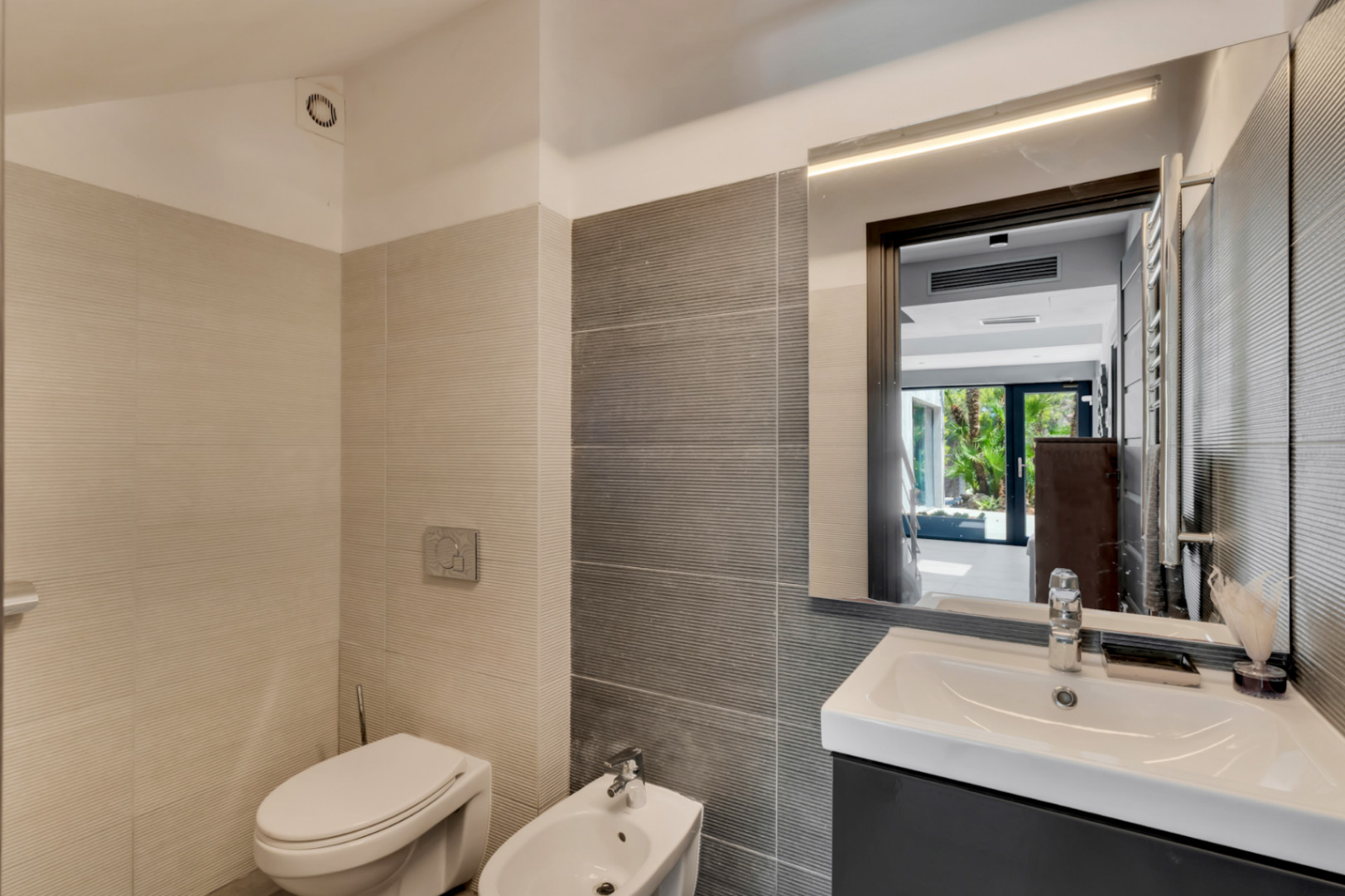 Salle de bain moderne avec carrelage texturé bicolore, toilettes, bidet et meuble-lavabo sombre. Le miroir éclairé reflète un jardin extérieur lumineux.