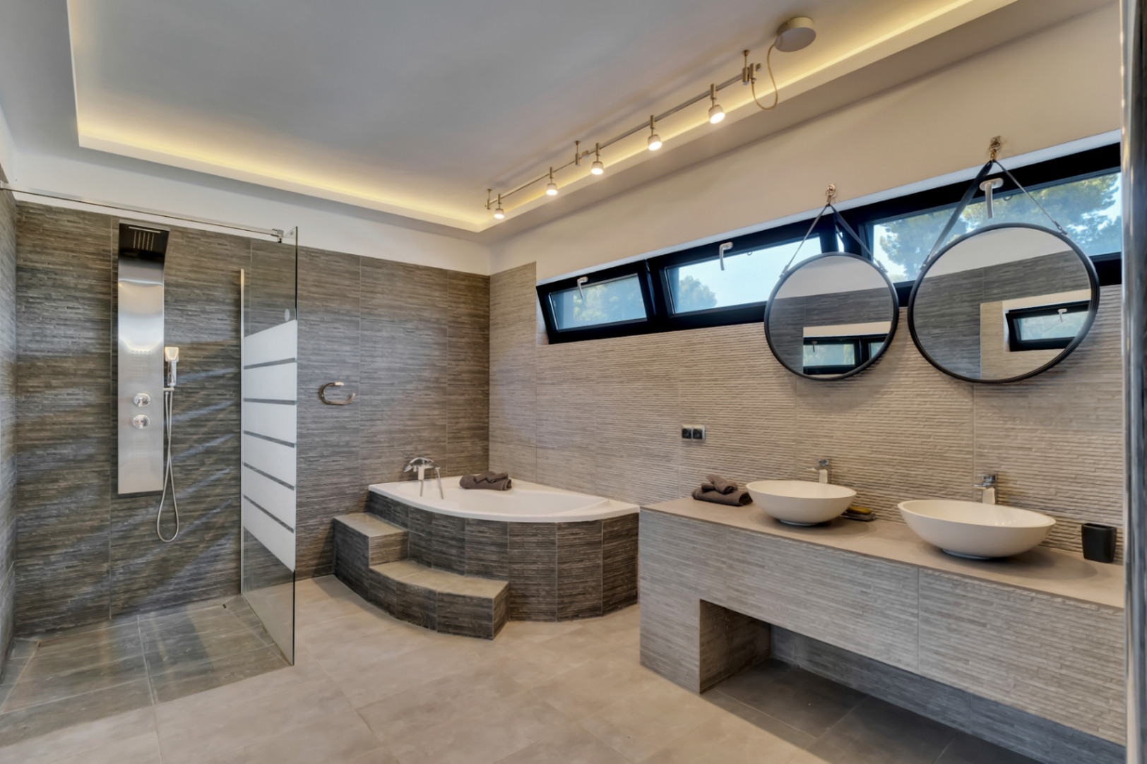 Salle de bain moderne avec douche à l'italienne, baignoire d'angle à marches et double vasque. Carreaux gris texturés, miroirs ronds suspendus et lumière naturelle des fenêtres.