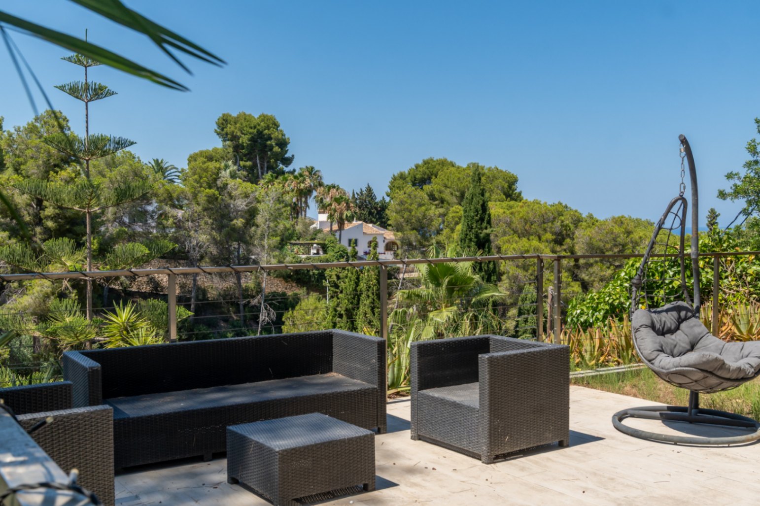 Terrasse extérieure aménagée avec mobilier en rotin foncé et fauteuil suspendu, offrant des vues panoramiques sur la végétation luxuriante et une maison lointaine.