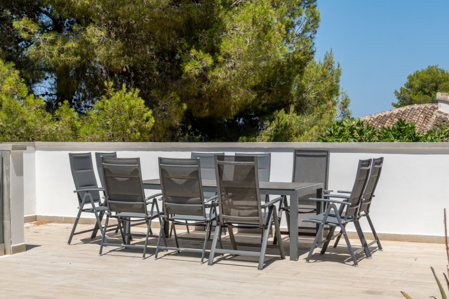 Terrasse extérieure carrelée avec table à manger moderne et huit chaises, délimitée par un mur blanc, offrant une vue sur la végétation luxuriante et les toits en tuiles traditionnelles.