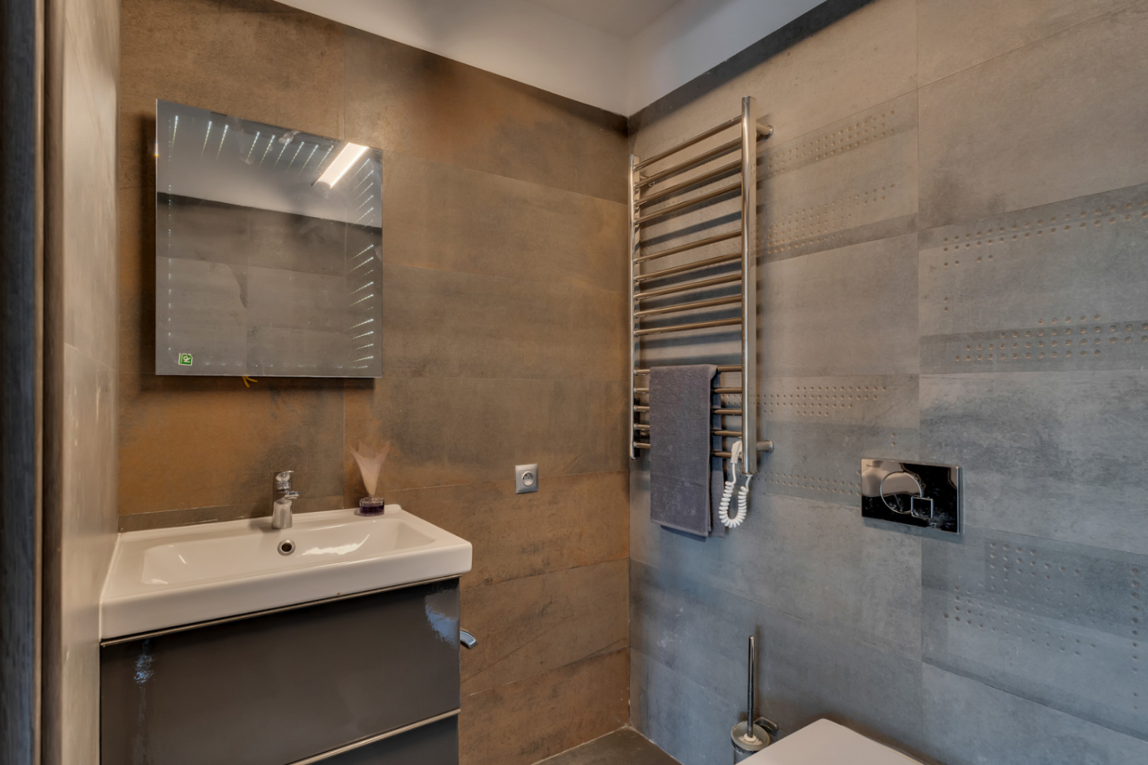 Toilette d'invités avec lavabo suspendu sur meuble gris foncé, miroir lumineux et sèche-serviettes chromé. Les carreaux muraux texturés gris et rouille apportent une touche contemporaine.