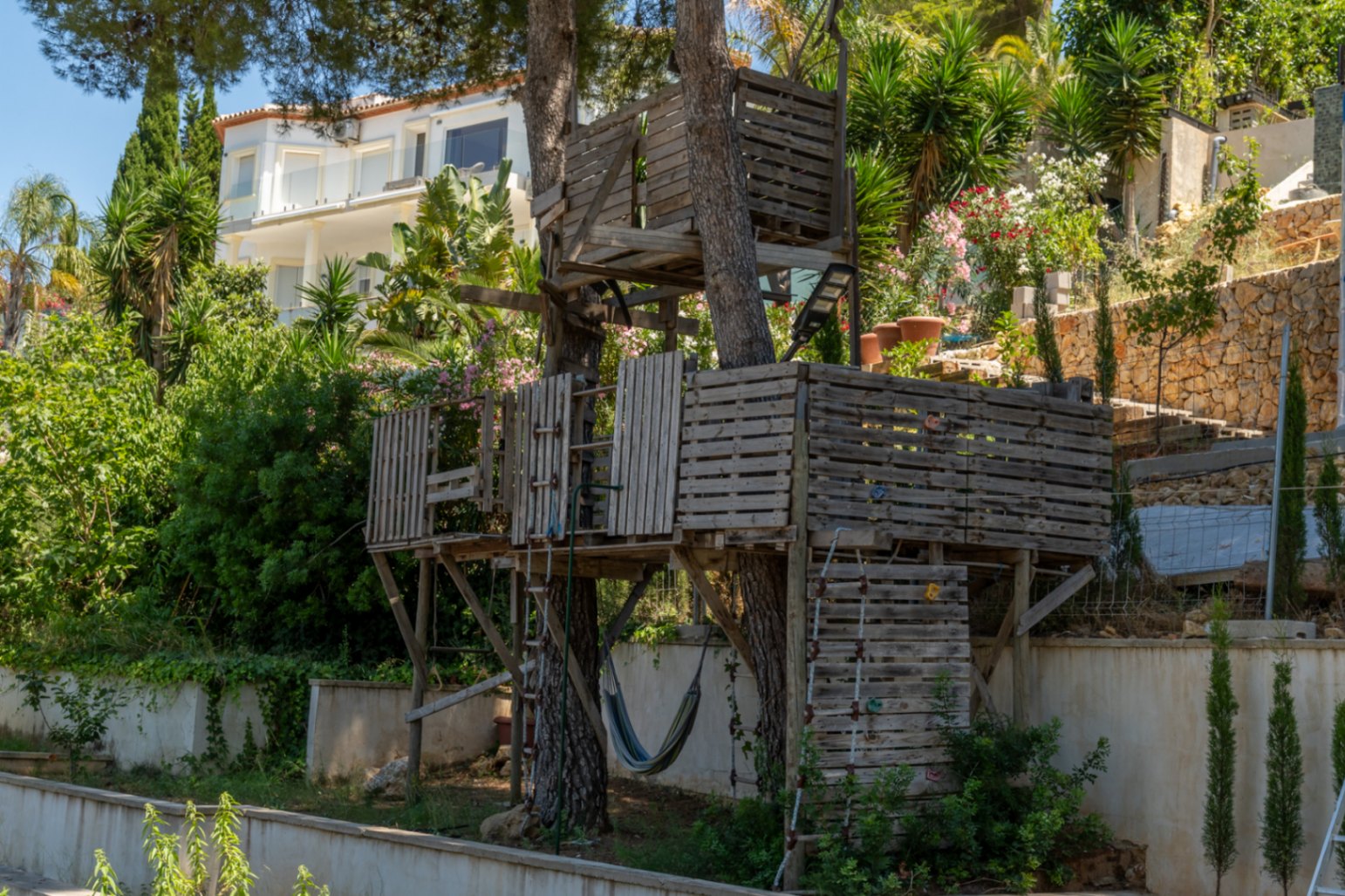 Une cabane dans les arbres rustique à plusieurs niveaux, construite avec des palettes en bois, dotée d'échelles et d'un hamac, entourée de verdure luxuriante avec une villa moderne en arrière-plan.