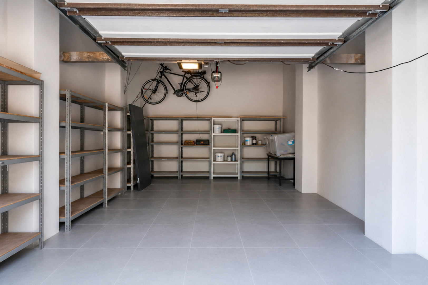 Vaste garage avec carrelage gris durable, étagères métalliques pour rangement organisé et support mural pour vélo. La structure apparente du plafond ajoute une touche pratique.