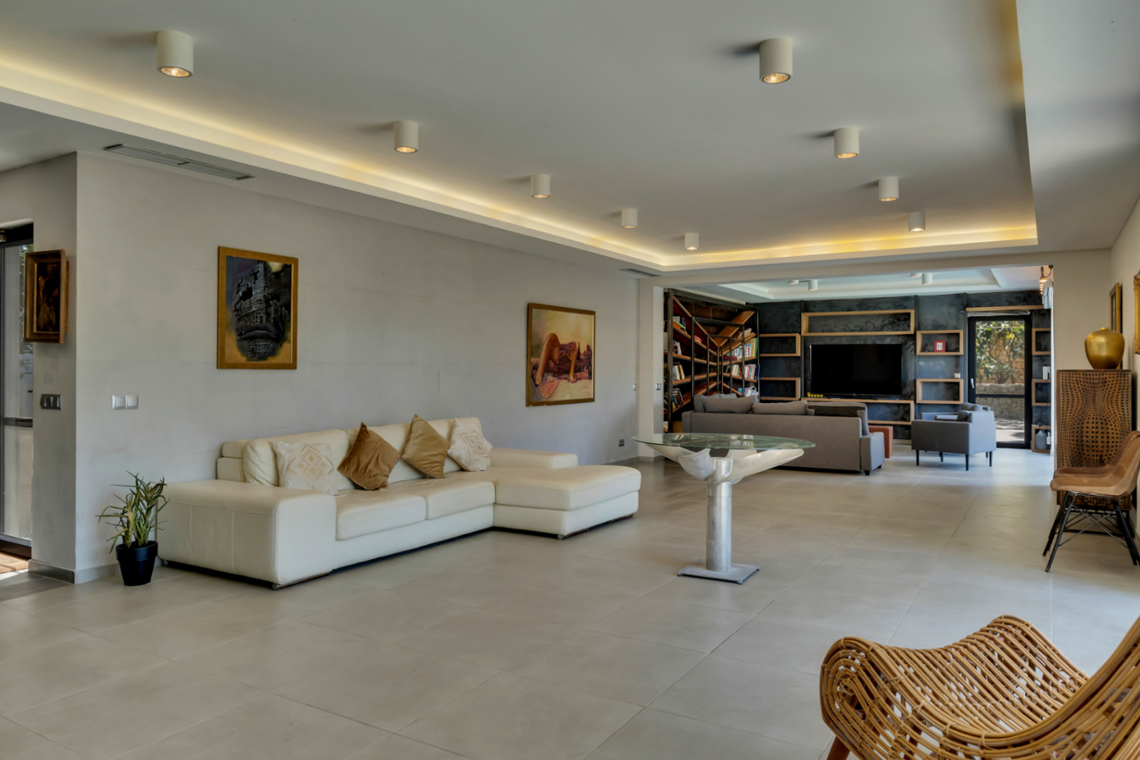 Vaste salon avec canapé d'angle blanc et table design, s'ouvrant sur un espace média et bibliothèque avec mur d'accent sombre et étagères en bois.