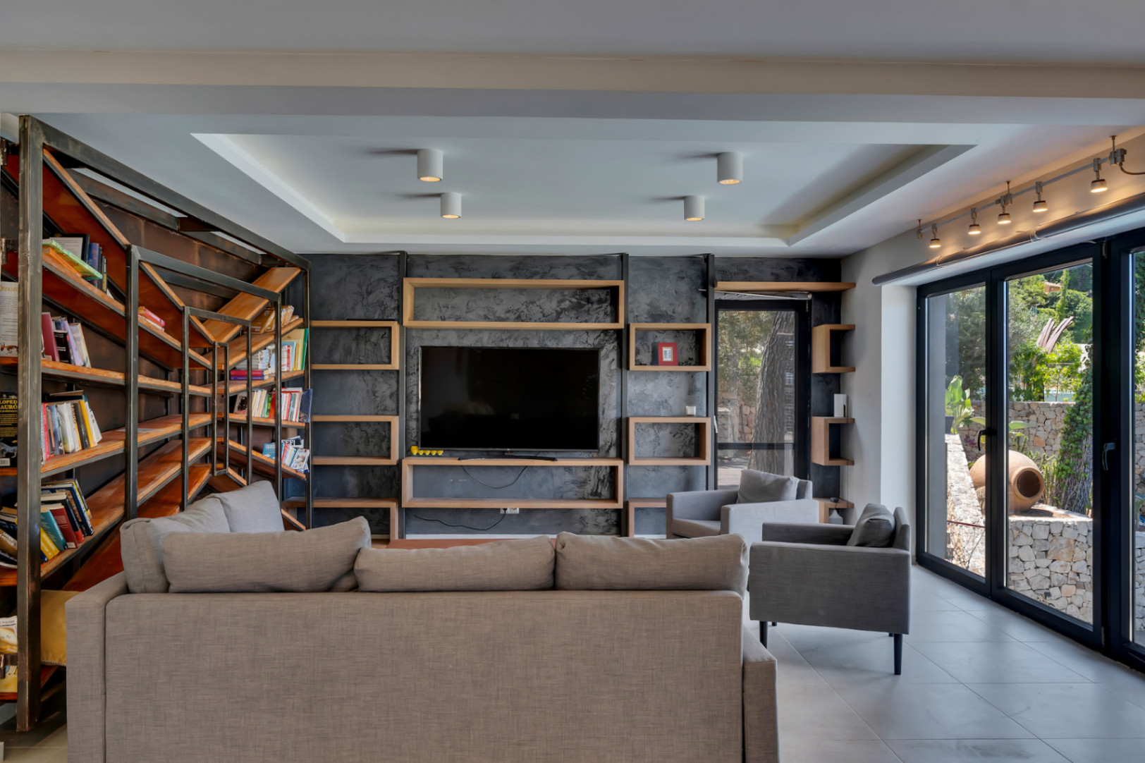 Vaste salon avec mur média texturé sombre et étagères en bois intégrées, complété par une bibliothèque industrielle et de grandes baies vitrées sur le jardin méditerranéen.