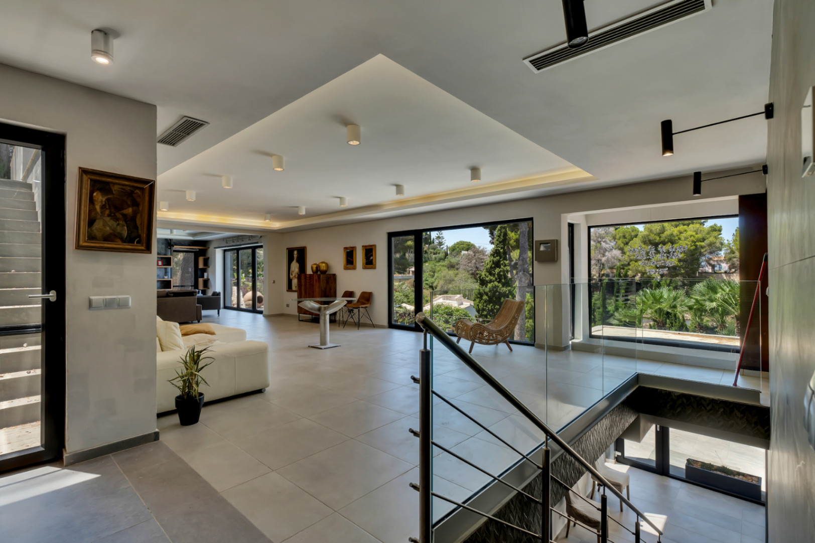 Vaste salon ouvert avec carrelage gris clair, de grandes baies vitrées offrant des vues sur la verdure luxuriante et un éclairage encastré moderne. La balustrade en verre de l'escalier apporte une touche contemporaine.