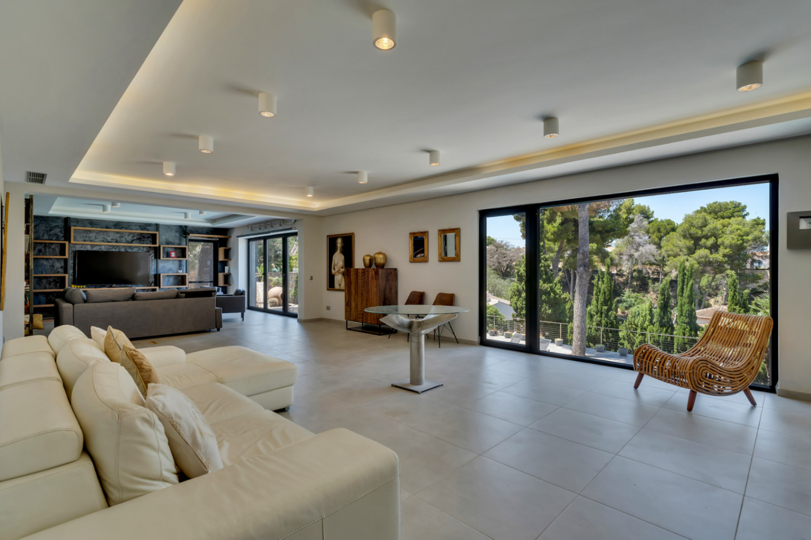 Vaste salon ouvert avec de grandes baies vitrées, offrant une vue sur la végétation luxuriante et une lumière naturelle abondante. Un canapé d'angle blanc et un éclairage encastré moderne caractérisent l'espace.