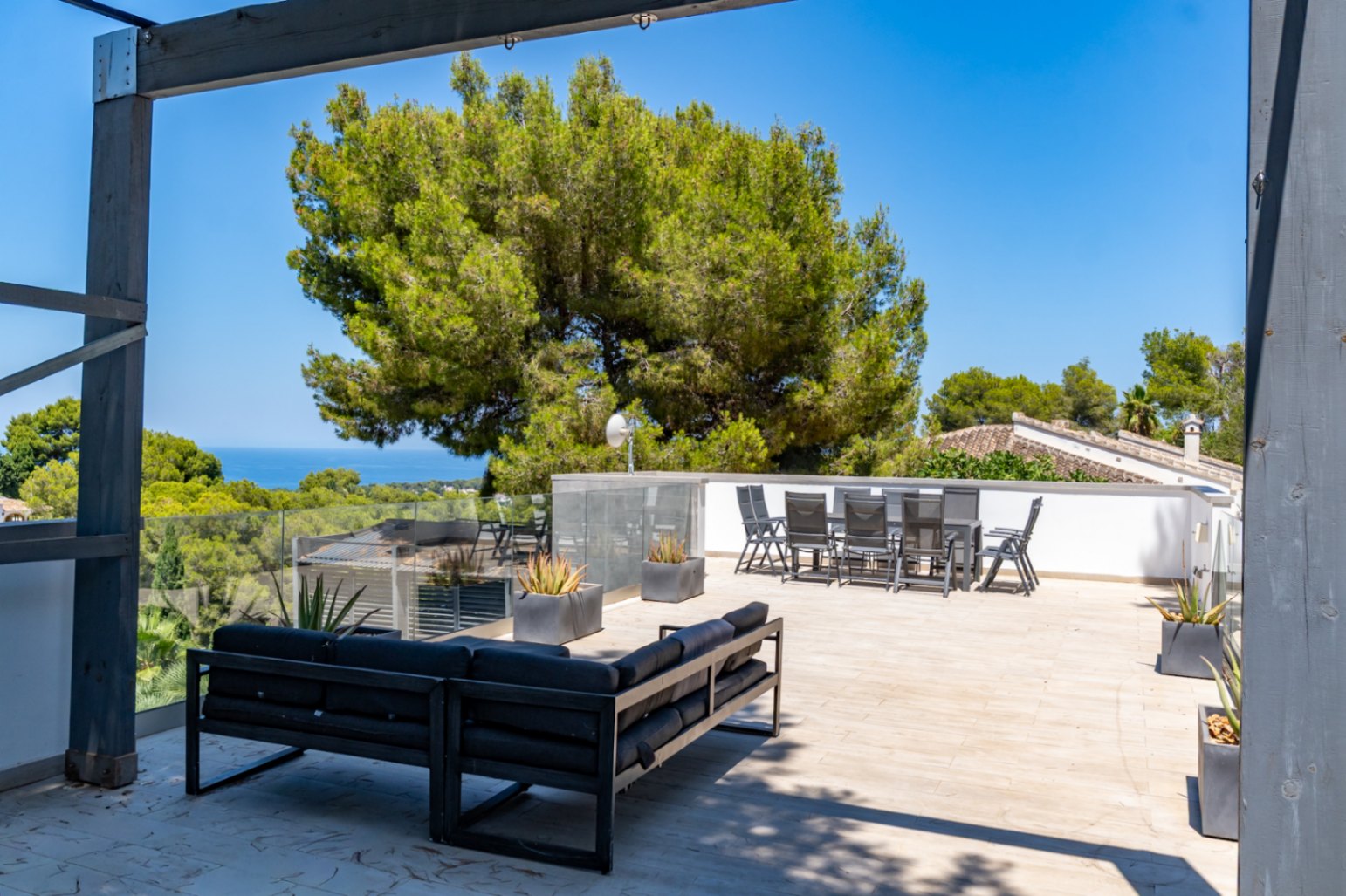 Vaste solarium sur le toit avec canapé extérieur moderne et ensemble repas, balustrades en verre et vues panoramiques sur la mer encadrées par les pins méditerranéens.