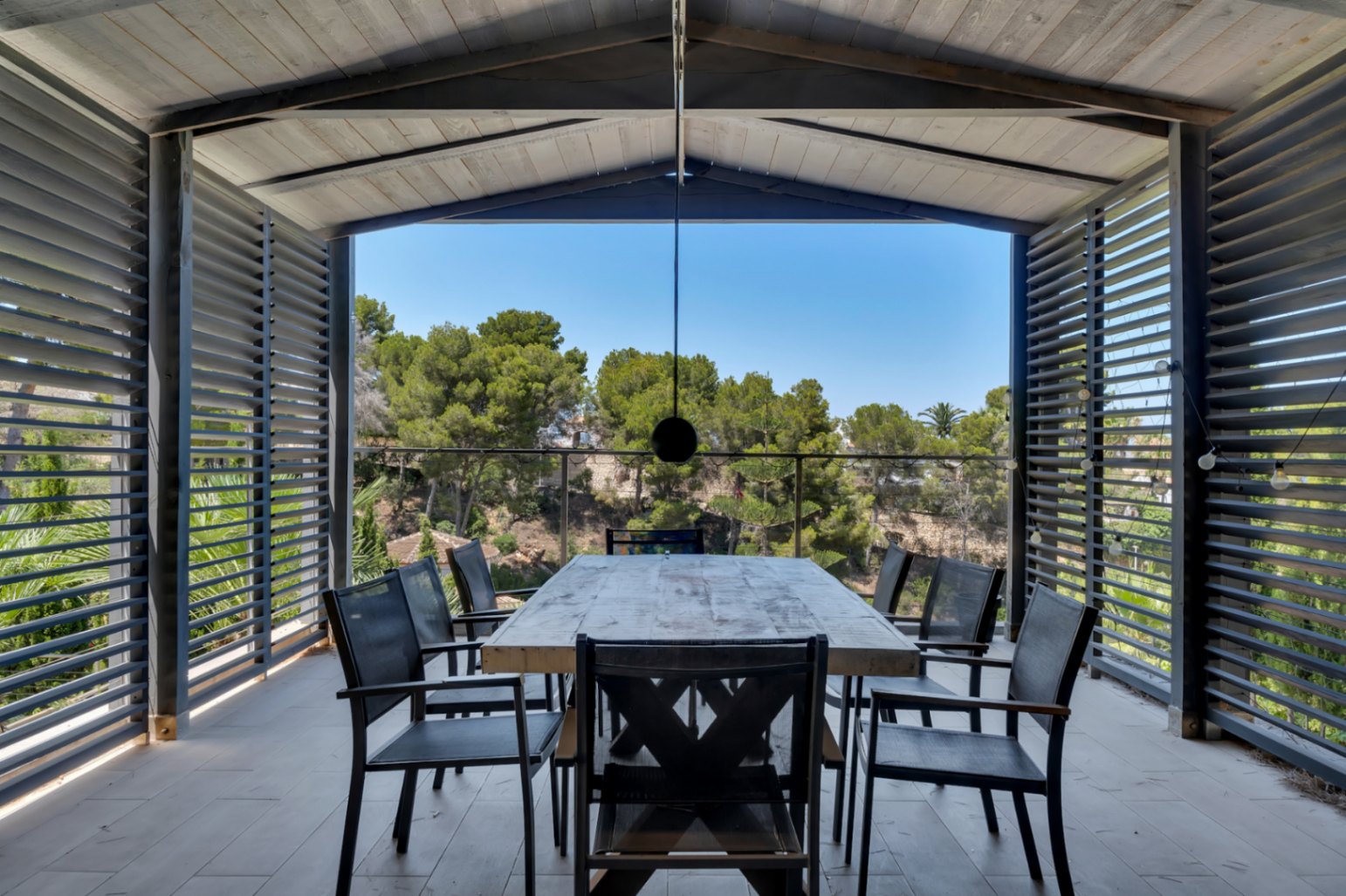 Vaste terrasse couverte avec table à manger en bois rustique et chaises sombres, encadrée de panneaux à persiennes offrant intimité et vue dégagée sur la végétation méditerranéenne.