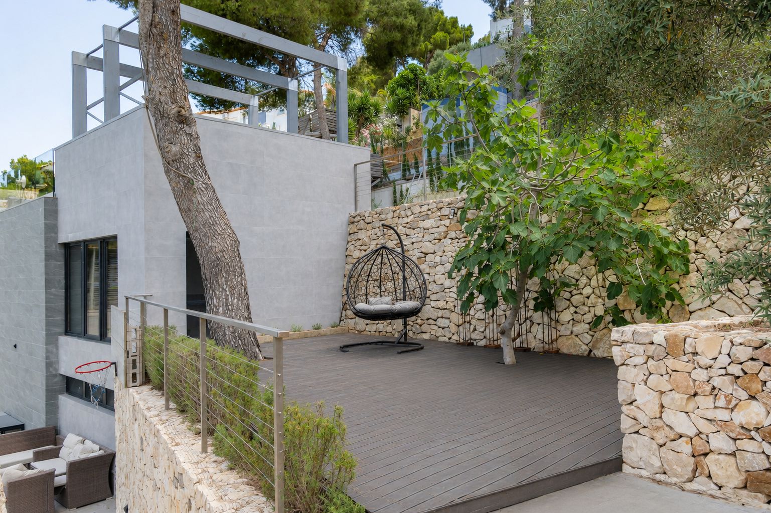 Vaste terrasse extérieure en composite foncé, agrémentée d'un fauteuil suspendu et encadrée de murs en pierre naturelle. La végétation méditerranéenne et l'architecture moderne se complètent harmonieusement.