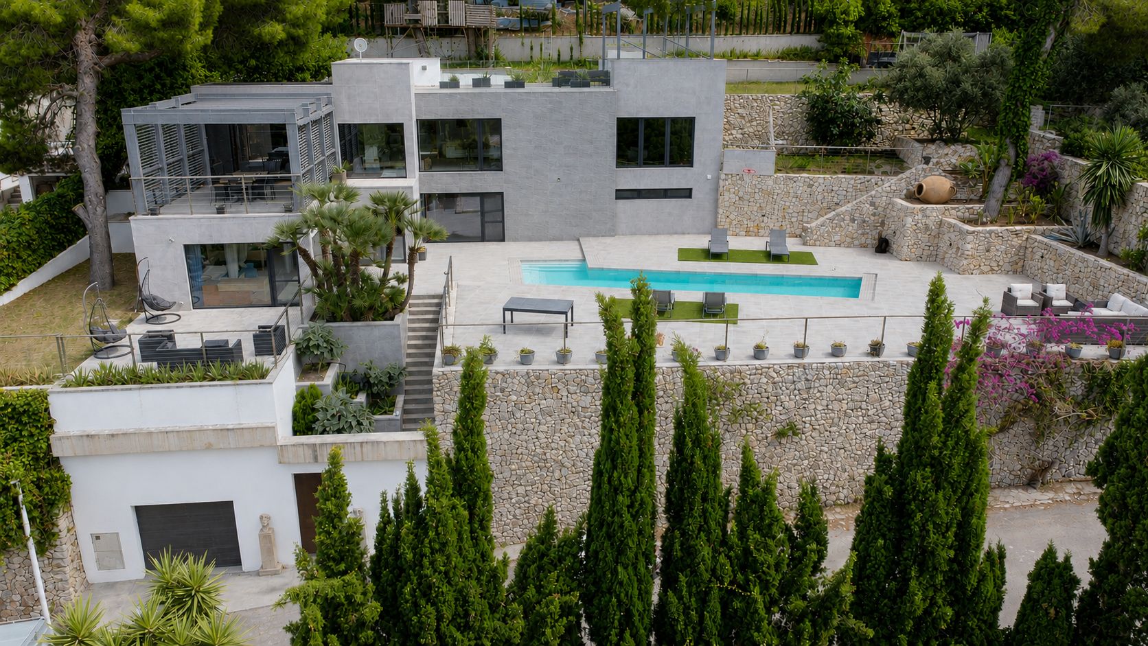 Vue aérienne de la villa moderne avec piscine rectangulaire et terrasses étagées. La façade grise, les jardins en restanques de pierre et les vastes espaces extérieurs sont visibles.