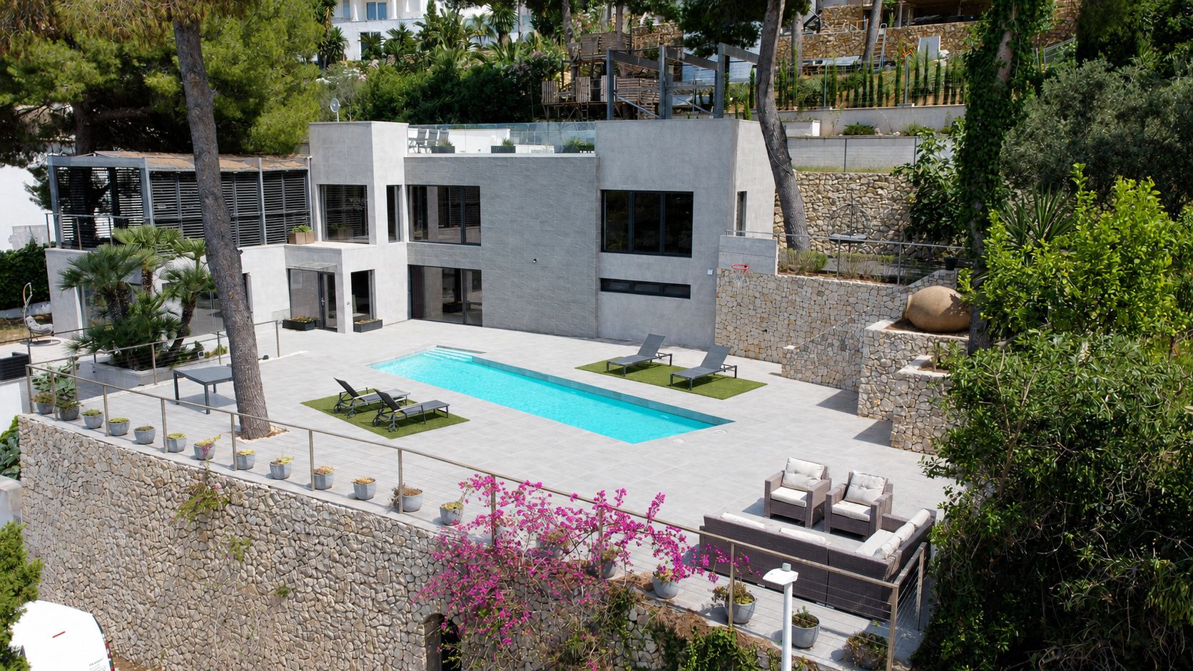 Vue aérienne de la villa moderne, avec sa façade grise épurée, sa piscine privée rectangulaire et ses vastes terrasses carrelées. Murs en pierre et végétation méditerranéenne luxuriante.