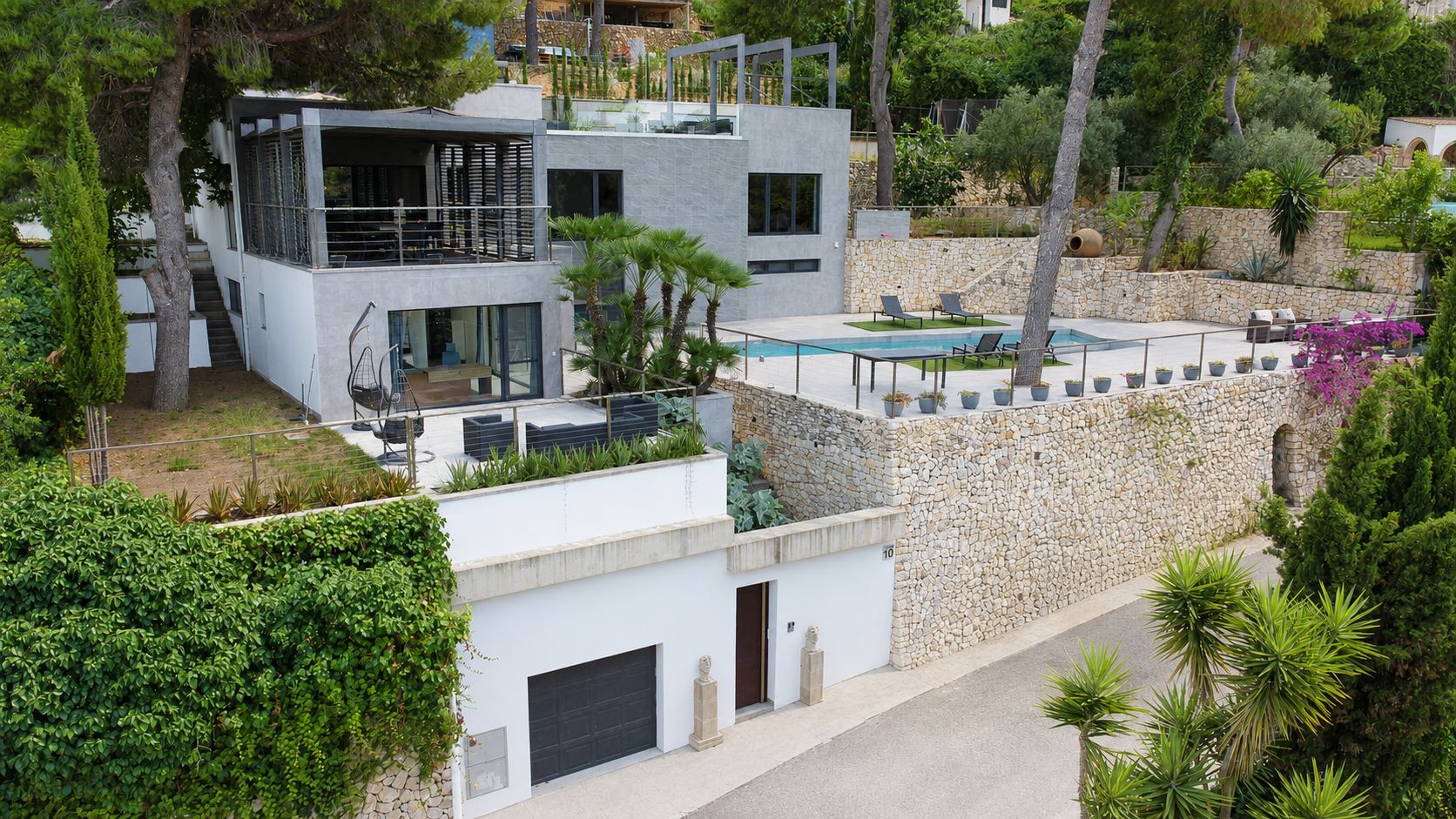 Vue aérienne de la villa moderne, intégrant une piscine privée, des terrasses aménagées et des murs en pierre naturelle, avec un garage au niveau de la rue.