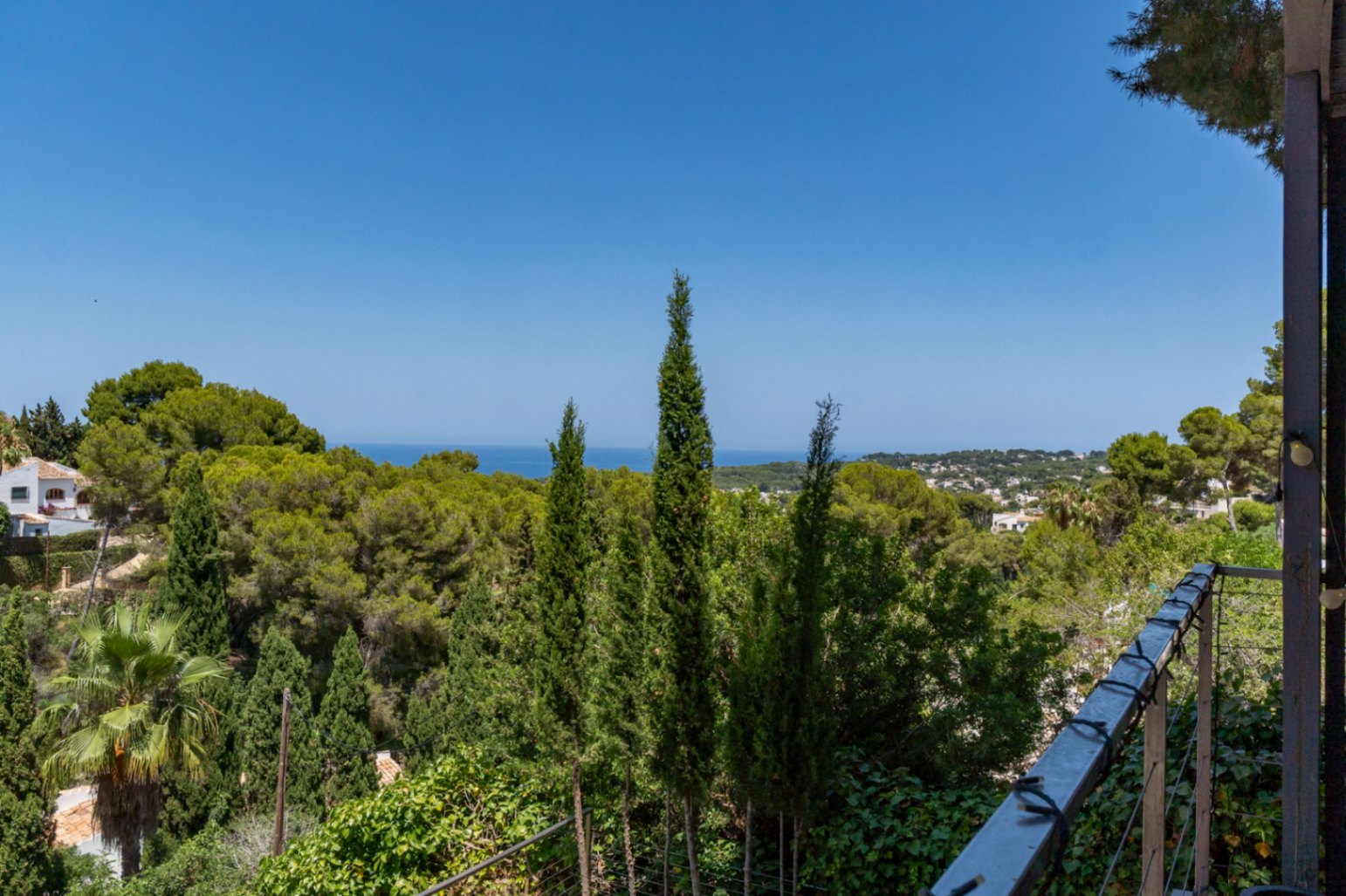 Vue depuis la terrasse sur une végétation méditerranéenne dense, des cyprès élancés, la mer lointaine et des zones résidentielles de Jávea sous un ciel azur.