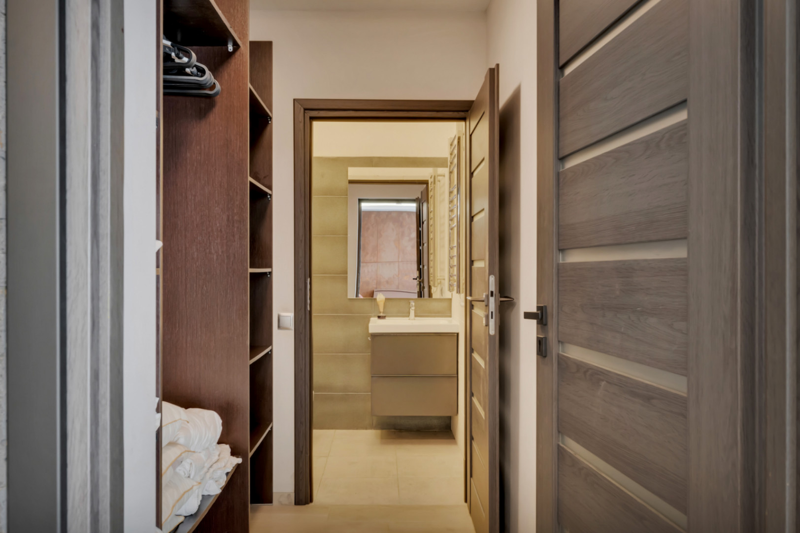 Vue d'un couloir vers une salle de bain moderne avec meuble-lavabo, miroir et murs carrelés. Une armoire ouverte en bois foncé avec étagères est visible à gauche.