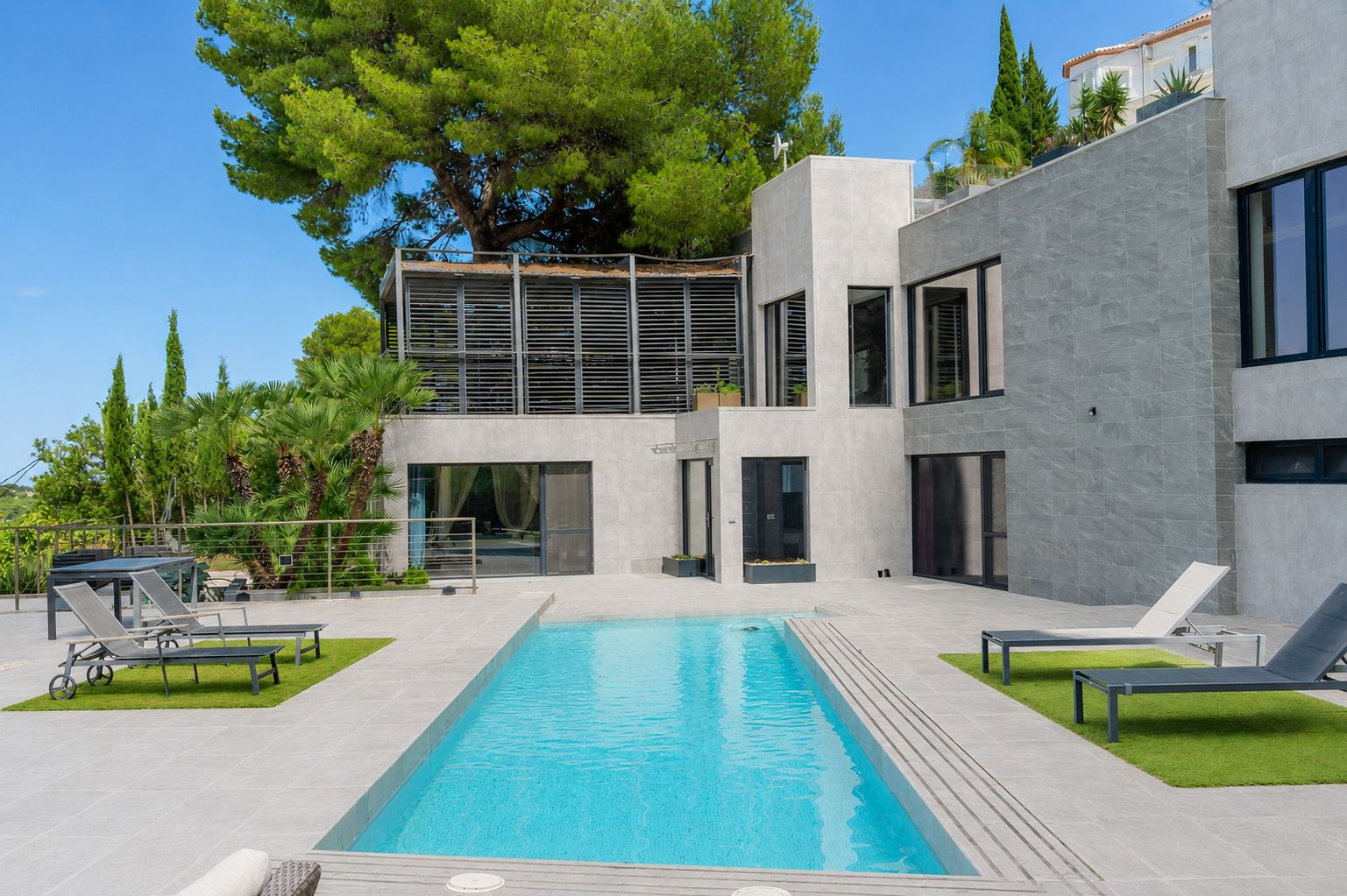 Vue extérieure de la villa avec piscine privée rectangulaire, terrasses carrelées grises et zones de gazon artificiel. L'architecture moderne et une terrasse supérieure à lames se distinguent.