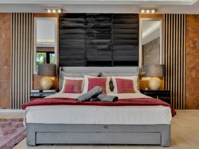 Chambre principale avec lit tapissé gris, tête de lit noire texturée, miroirs et panneaux muraux uniques en bois, métal vieilli et pierre. L'éclairage sur mesure crée une ambiance.