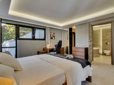 Chambre principale lumineuse avec lit double, bureau en bois et grandes fenêtres. Comprend une armoire à portes miroir, une commode et un accès direct à une salle de bain privative carrelée.