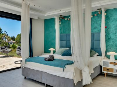 Chambre spacieuse avec lit à baldaquin, tête de lit bleue capitonnée, mur d'accent turquoise texturé et accès direct à une terrasse extérieure avec fauteuils suspendus.