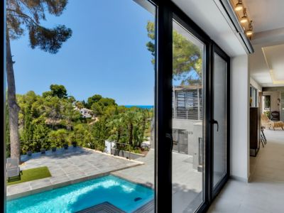 De larges baies vitrées noires relient l'intérieur moderne à la piscine privée, la terrasse carrelée et le paysage méditerranéen luxuriant avec vue sur la mer lointaine.
