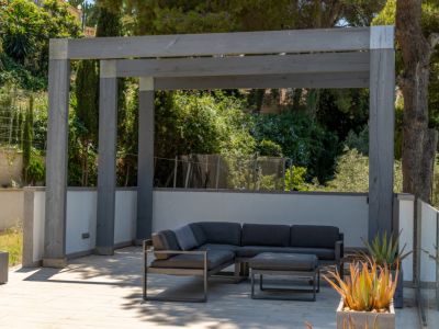 Espace salon extérieur moderne sous une pergola en bois gris, avec canapé d'angle et pouf sur un sol pavé clair, entouré de verdure méditerranéenne luxuriante et de plantes grasses.