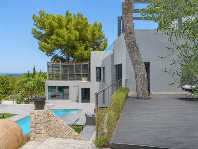 Extérieur de villa moderne à plusieurs niveaux, avec façades grises, piscine privée et terrasse en composite intégrant un arbre. Vues mer lointaines à travers la végétation méditerranéenne.
