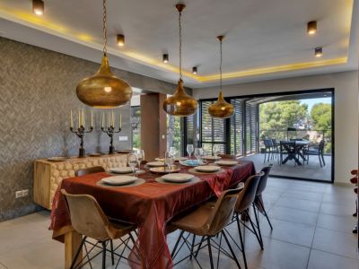 Grande salle à manger avec table en bois et chaises contemporaines, éclairée par trois suspensions dorées et un éclairage indirect. Le mur texturé et le buffet sculpté s'ouvrent sur une terrasse couverte.