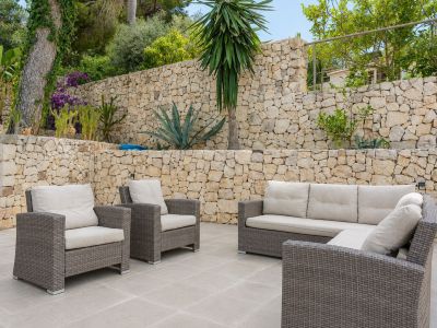Grande terrasse extérieure avec mobilier en rotin et coussins clairs, encadrée par des murs en pierre sèche étagés et une végétation méditerranéenne luxuriante.