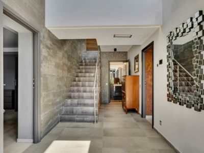 Hall d'entrée spacieux avec un escalier moderne revêtu de carreaux effet béton et une rampe en acier inoxydable. Le sol est carrelé de gris et un miroir modulaire décore le mur.
