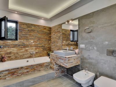 Salle de bain moderne avec baignoire et meuble-lavabo suspendu, tous deux revêtus de pierre empilée multicolore. De grands carreaux gris habillent murs et sol, éclairage encastré.