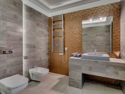 Salle de bain moderne avec carrelage mural gris texturé, un mur d'accent 3D géométrique bronze et un meuble-lavabo suspendu. Comprend toilettes et bidet, ainsi qu'un sèche-serviettes.