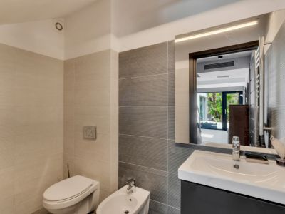 Salle de bain moderne avec carrelage texturé bicolore, toilettes, bidet et meuble-lavabo sombre. Le miroir éclairé reflète un jardin extérieur lumineux.