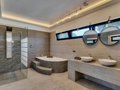 Salle de bain moderne avec douche à l'italienne, baignoire d'angle à marches et double vasque. Carreaux gris texturés, miroirs ronds suspendus et lumière naturelle des fenêtres.