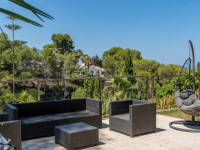 Terrasse extérieure aménagée avec mobilier en rotin foncé et fauteuil suspendu, offrant des vues panoramiques sur la végétation luxuriante et une maison lointaine.