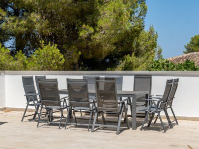 Terrasse extérieure carrelée avec table à manger moderne et huit chaises, délimitée par un mur blanc, offrant une vue sur la végétation luxuriante et les toits en tuiles traditionnelles.