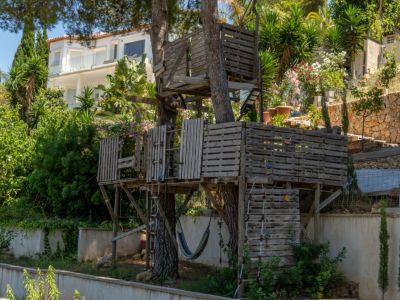 Une cabane dans les arbres rustique à plusieurs niveaux, construite avec des palettes en bois, dotée d'échelles et d'un hamac, entourée de verdure luxuriante avec une villa moderne en arrière-plan.