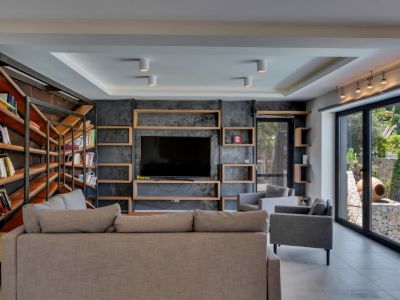 Vaste salon avec mur média texturé sombre et étagères en bois intégrées, complété par une bibliothèque industrielle et de grandes baies vitrées sur le jardin méditerranéen.