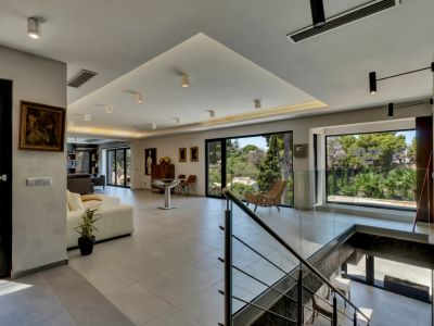 Vaste salon ouvert avec carrelage gris clair, de grandes baies vitrées offrant des vues sur la verdure luxuriante et un éclairage encastré moderne. La balustrade en verre de l'escalier apporte une touche contemporaine.