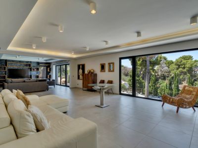 Vaste salon ouvert avec de grandes baies vitrées, offrant une vue sur la végétation luxuriante et une lumière naturelle abondante. Un canapé d'angle blanc et un éclairage encastré moderne caractérisent l'espace.
