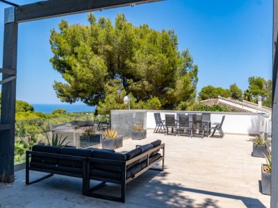 Vaste solarium sur le toit avec canapé extérieur moderne et ensemble repas, balustrades en verre et vues panoramiques sur la mer encadrées par les pins méditerranéens.